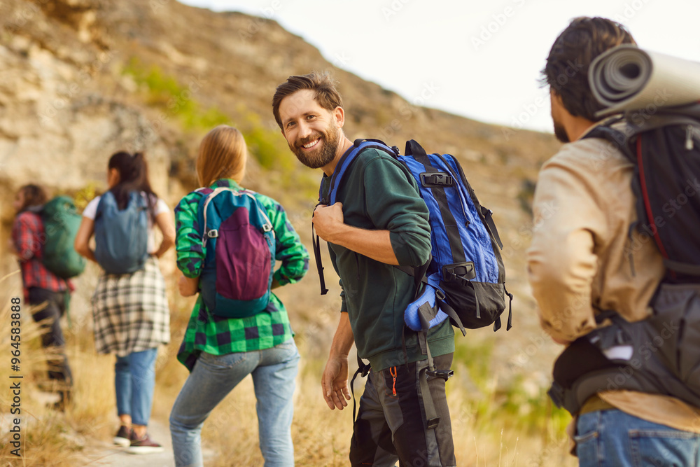 Portrait of a happy man hiker, wearing backpack enjoying a hike with a group of friends. This team is on a trip, making the most of vacation or weekend by spending time together in nature. Portrait of a happy man hiker, wearing backpack enjoying a hike with a group of friends. This team is on a trip, making the most of vacation or weekend by spending time together in nature.