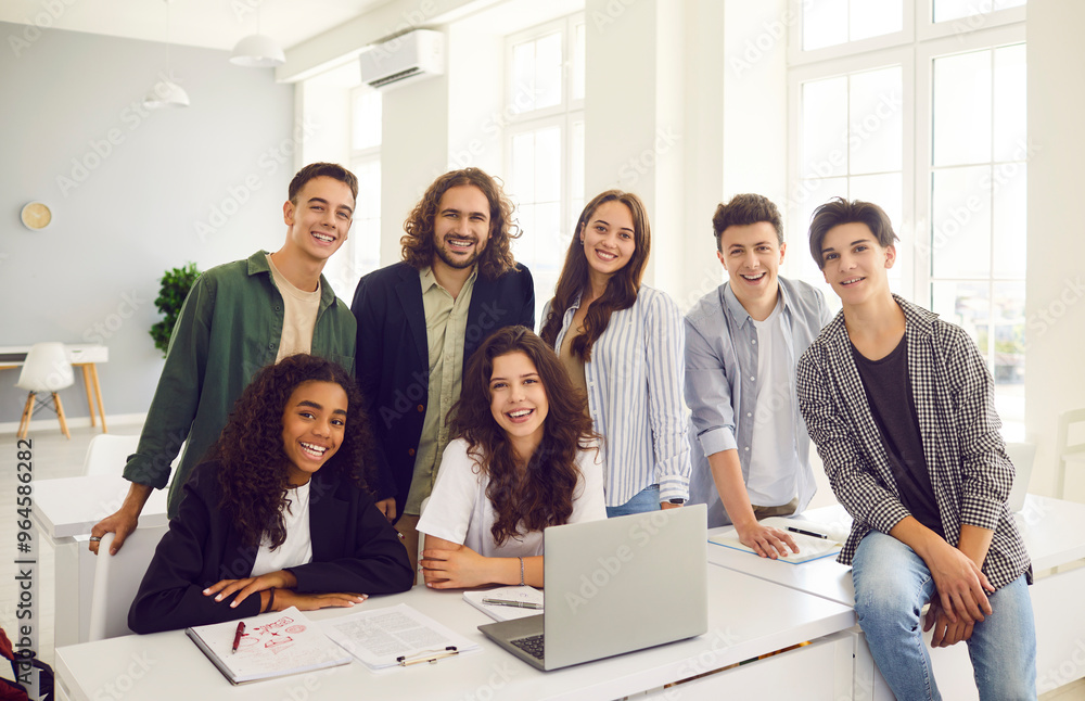 Group of high school students and classmates sitting at the desk with male friendly smiling teacher in modern classroom with laptop and looking at the camera. Education and back to school concept. Group of high school students and classmates sitting at the desk with male friendly smiling teacher in modern classroom with laptop and looking at the camera. Education and back to school concept.