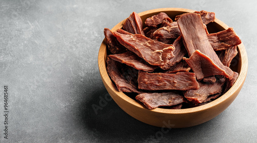 Wooden bowl filled with pieces of beef jerky on a dark textured surface. The dried meat is arranged in an overhead view, showcasing its texture and color.