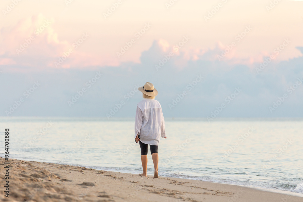 Woman wearing white shirt and straw hat walking on beach at sunset Woman wearing white shirt and straw hat walking on beach at sunset