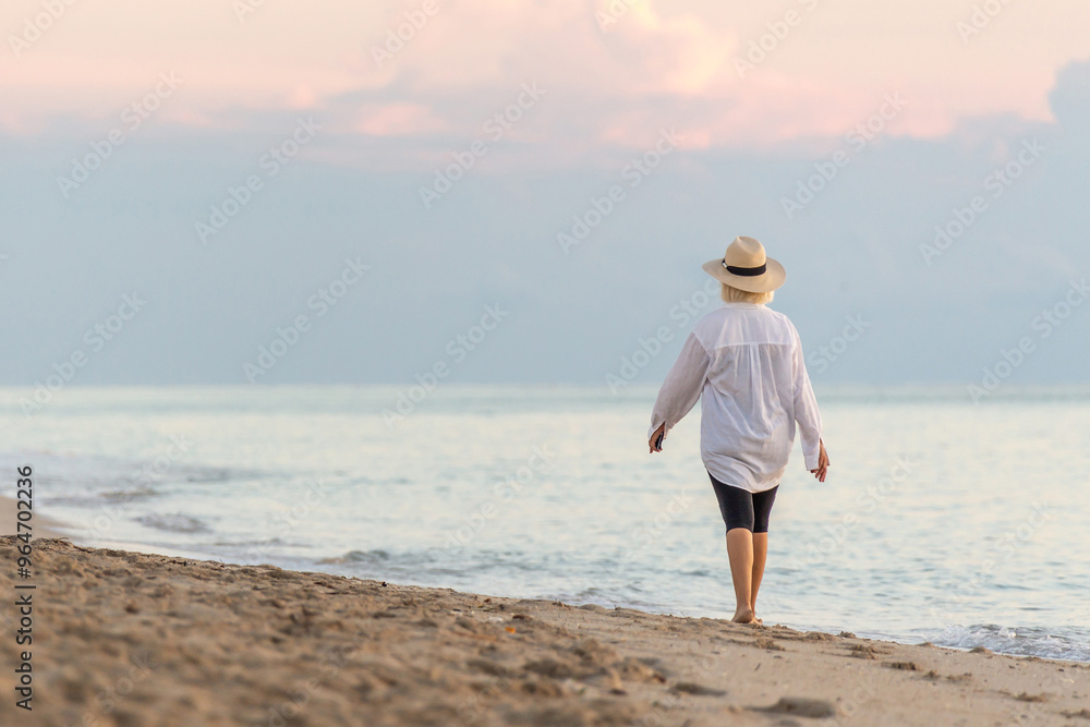 Woman walking along beach at sunset holding cell phone and wearing straw hat Woman walking along beach at sunset holding cell phone and wearing straw hat