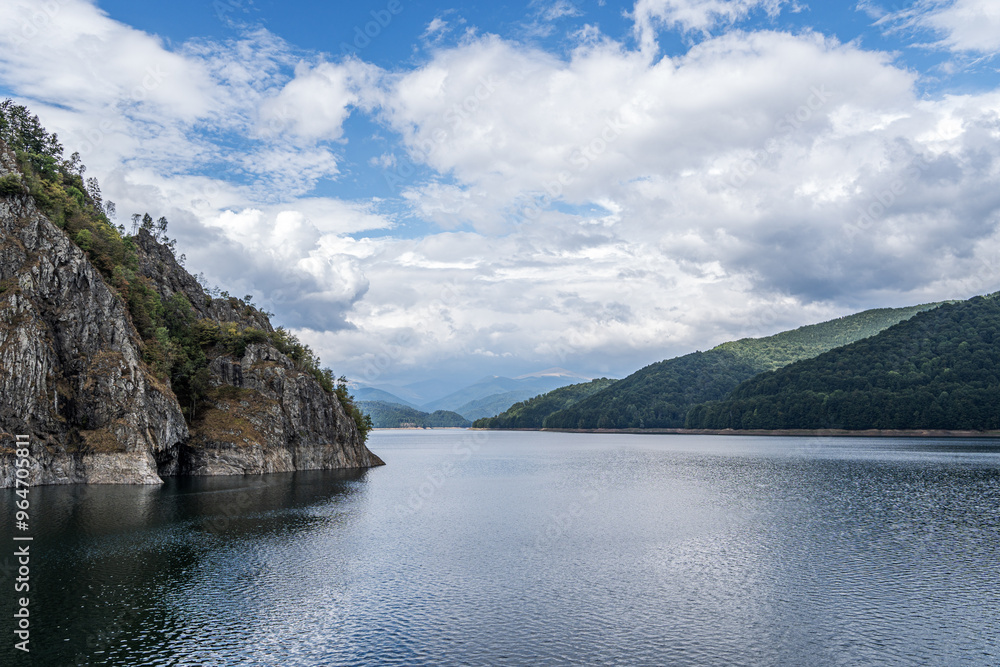A serene lake is surrounded by rocky cliffs and lush green hills under a partly cloudy sky. The peaceful water reflects the natural beauty, while distant mountains add depth to the scenic view. A serene lake is surrounded by rocky cliffs and lush green hills under a partly cloudy sky. The peaceful water reflects the natural beauty, while distant mountains add depth to the scenic view.