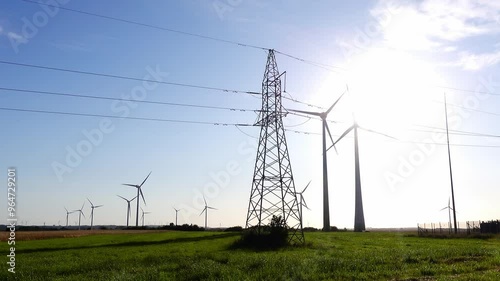 Fast spinning rotors of wind turbines on the farmlands. High voltage pylon in the foreground. Shot taken against the sun on a sunny day.