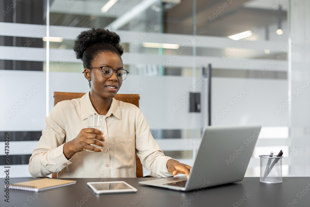 Confident African American businesswoman using laptop in office setting. She is holding glass of water, with tablet and phone visible on desk. Professional atmosphere suggests focus Confident African American businesswoman using laptop in office setting. She is holding glass of water, with tablet and phone visible on desk. Professional atmosphere suggests focus