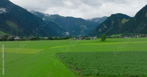 Take off shot with a drone flying over a field and reveal a city located in the Zillertal surrounded by mountains in the Alps, overcast summer day