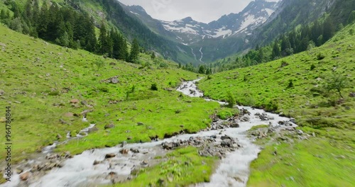 Aerial drone shot of a river in a valley near Zemmgrund surrounded by trees ,grass and mountains located in the Alps, the mountains in the background are partly covered with snow, summer day