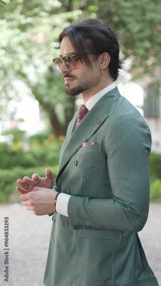 side view of sexy man with sunglasses rubbing palms, looking to side, adjusting green suit while standing in the nature side view of sexy man with sunglasses rubbing palms, looking to side, adjusting green suit while standing in the nature
