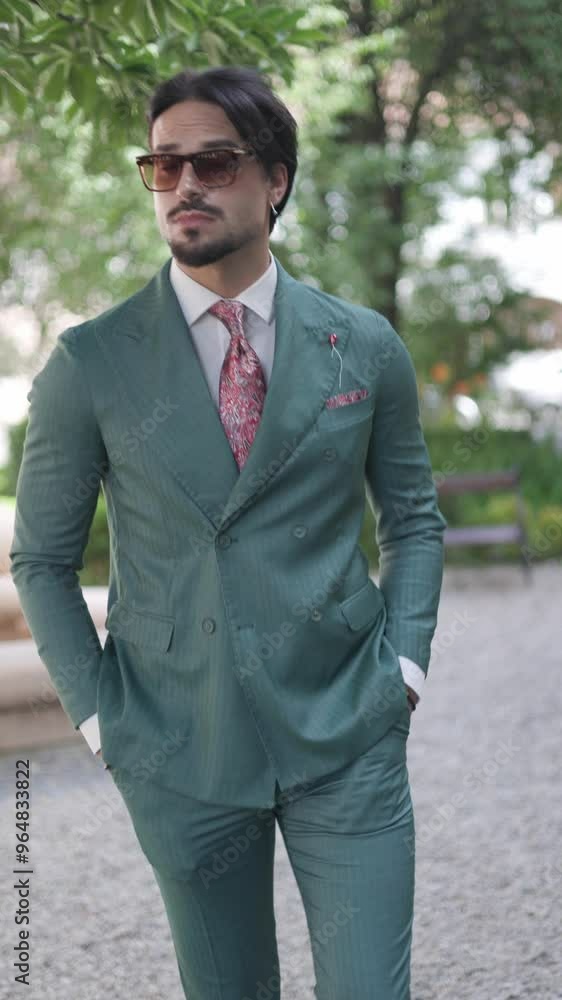 attractive young businessman in green suit with sunglasses walking with hands in pockets and looking around in the park attractive young businessman in green suit with sunglasses walking with hands in pockets and looking around in the park