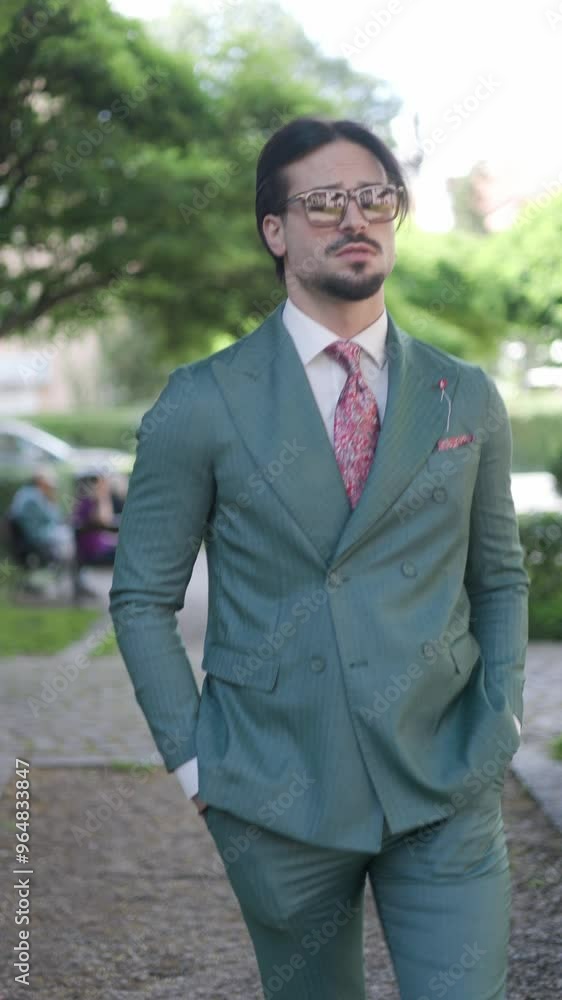 elegant fashion guy with sunglasses holding hands in pockets and walking while looking around in the park elegant fashion guy with sunglasses holding hands in pockets and walking while looking around in the park