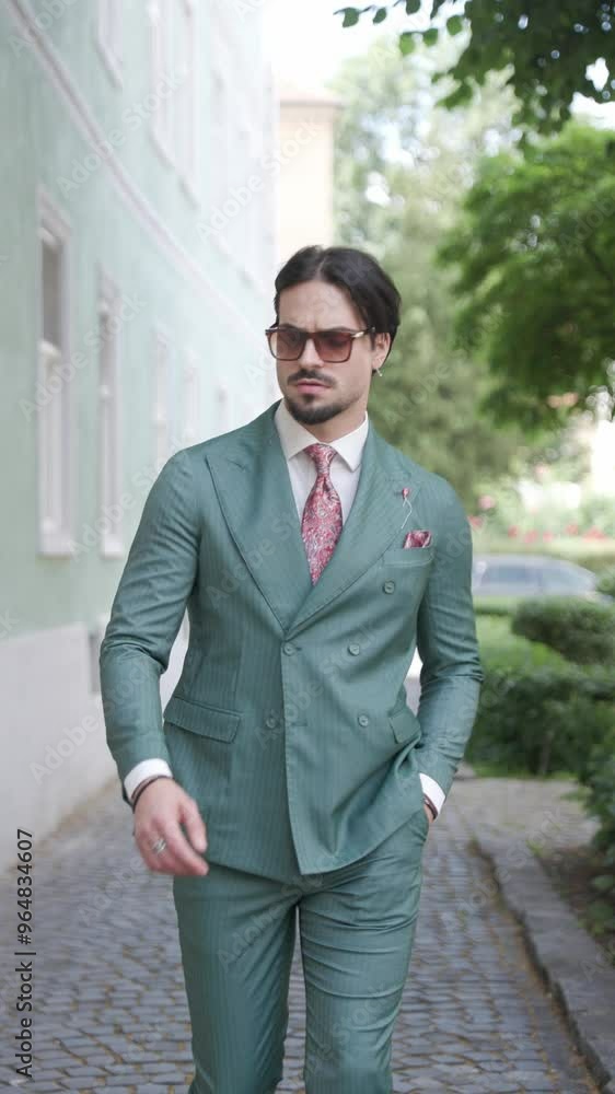 cool sexy businessman with sunglasses walking, looking to side and adjusting tie and green suit while being outside in the city cool sexy businessman with sunglasses walking, looking to side and adjusting tie and green suit while being outside in the city