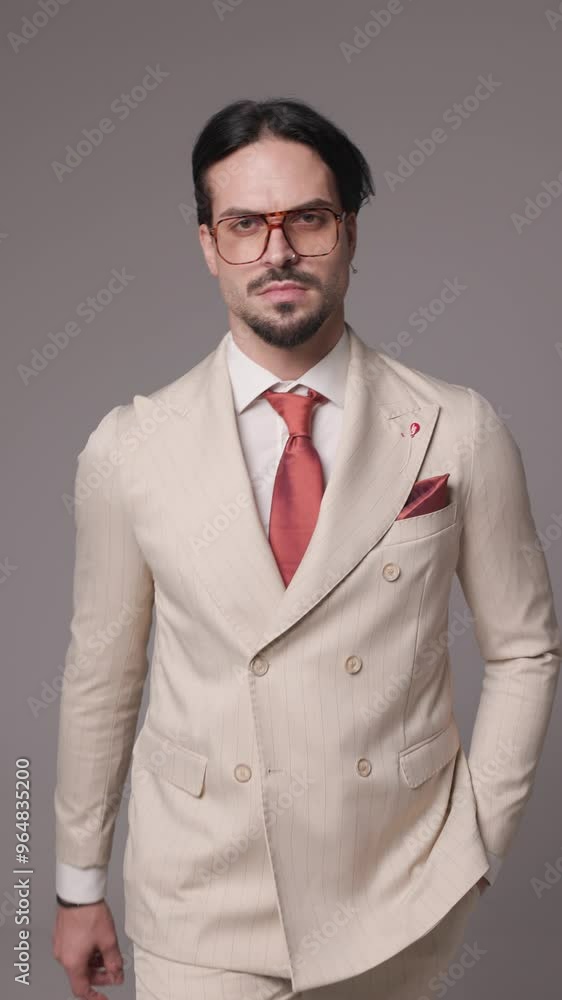 project video of confident young man with eyeglasses in beige suit stepping with hand in pocket and snapping fingers, looking down on grey background project video of confident young man with eyeglasses in beige suit stepping with hand in pocket and snapping fingers, looking down on grey background