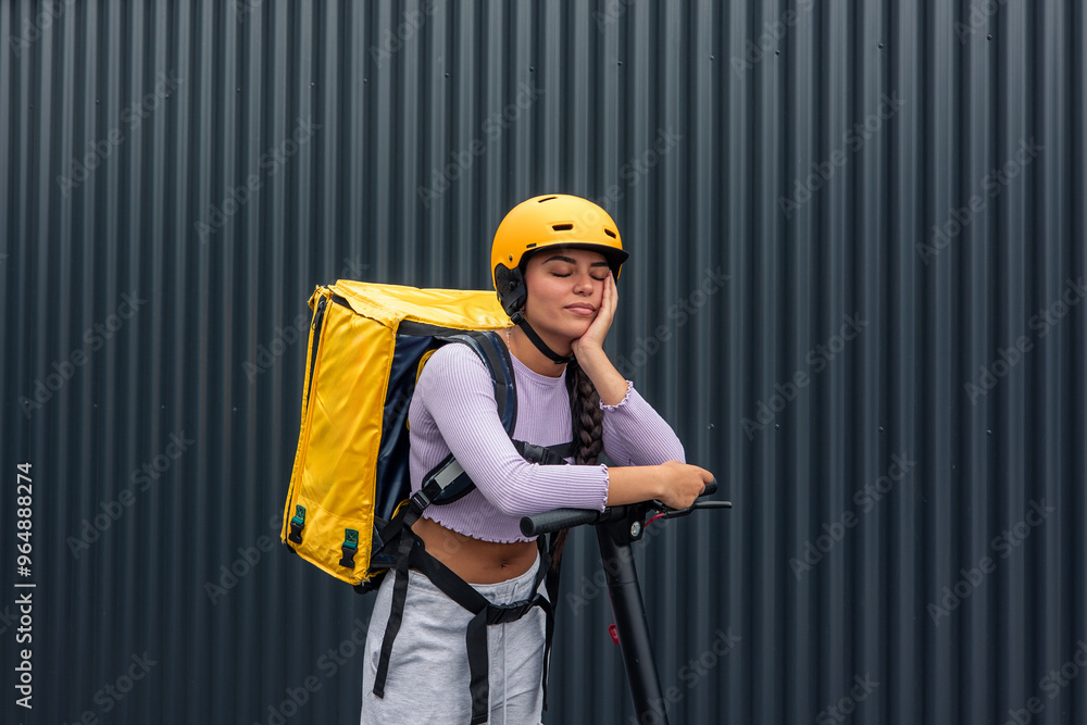 Bored Latin food courier with yellow backpack and helmet leaning on electric scooter Bored Latin food courier with yellow backpack and helmet leaning on electric scooter