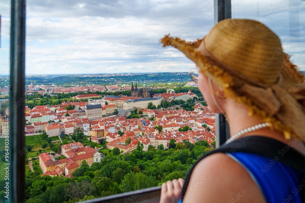 Fototapeta premium A female tourist wearing a stylish straw hat gazes at the stunning cityscape of Prague from the window of the iconic Prague Castle tower. The view showcases the rich history and beauty of the city.