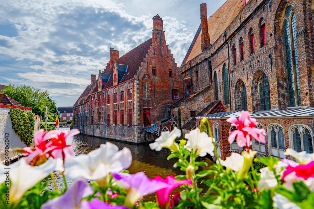 Naklejka premium On a lovely summer day in Bruges, Belgium, a charming canal is lined with vibrant flowers and historic buildings, making it an ideal spot for romantic getaways and relaxing vacations.