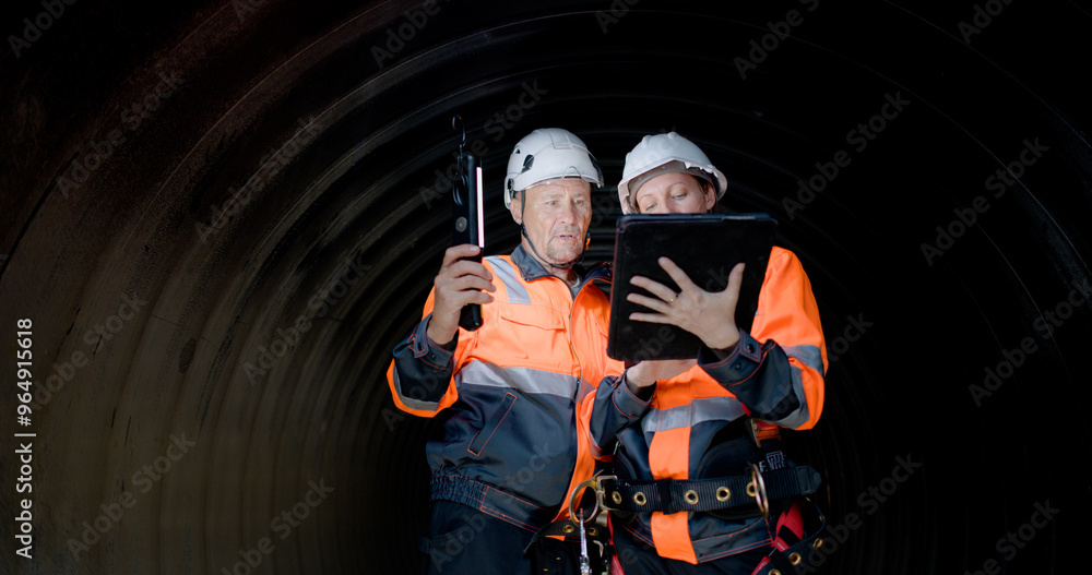 Fototapeta premium A couple of senior civil engineers are collaborating in the tunnel of a sewage drain, using a digital tablet to inspect the construction of a huge water pipe on a new highway road project.