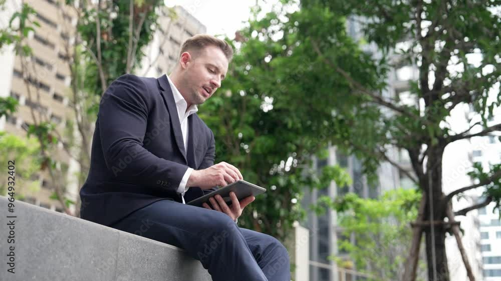 Side view of businessman sitting at park while writing on tablet by using tablet pencil. Low angle camera of smart project manager looking at data analysis while thinking about financial plan. Urbane Side view of businessman sitting at park while writing on tablet by using tablet pencil. Low angle camera of smart project manager looking at data analysis while thinking about financial plan. Urbane