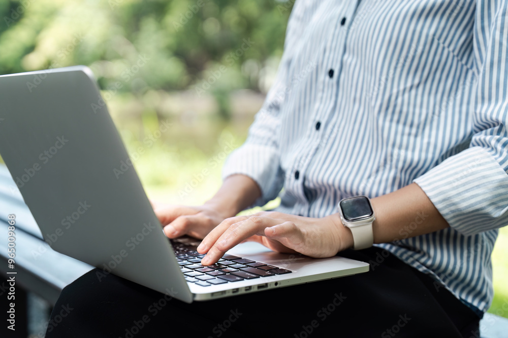 Fototapeta premium Close-up of Person Typing on Laptop Outdoors in a Park Setting, Wearing a Striped Shirt and Smartwatch