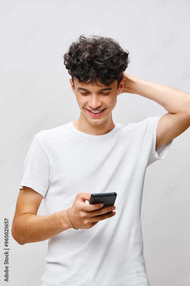 Young man using smartphone, smiling happily, wearing a plain white t shirt, standing against a light grey background, showcasing a sense of joy and connectivity Young man using smartphone, smiling happily, wearing a plain white t shirt, standing against a light grey background, showcasing a sense of joy and connectivity