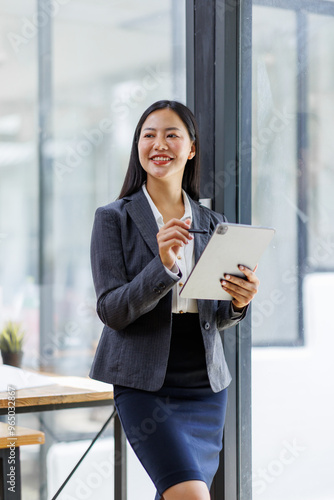 Modern business asian woman in formalwear using digital tablet while standing near wooden desk in an office interior in the office. business people concept.