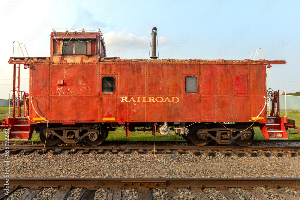 Naklejka premium Rusty Red Rail Car. An old train car sit abandoned on display in a small town. Yellow railroad painted on the sign as it sits idle on the tracks. A historic piece of transport and old time frieght