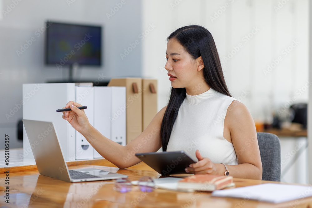 Positive young asian female freelancer in formal suit sitting at table with laptop and browsing tablet while working on project in creative workspace in daylight Positive young asian female freelancer in formal suit sitting at table with laptop and browsing tablet while working on project in creative workspace in daylight