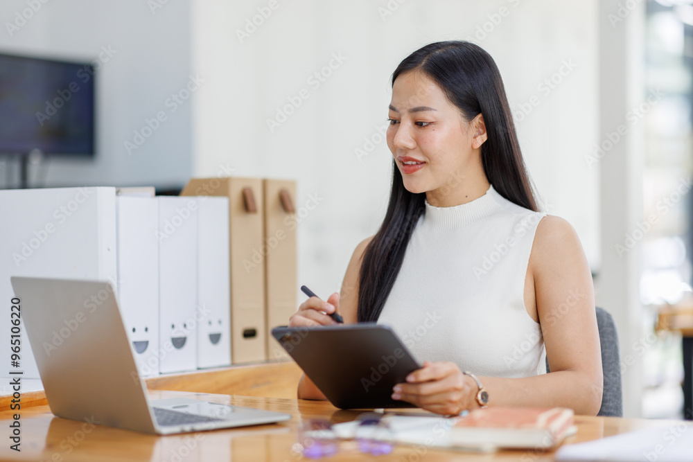 Positive young asian female freelancer in formal suit sitting at table with laptop and browsing tablet while working on project in creative workspace in daylight Positive young asian female freelancer in formal suit sitting at table with laptop and browsing tablet while working on project in creative workspace in daylight