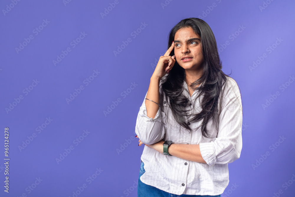 A young woman of Indian descent, wearing a white striped shirt and black smartwatch, poses thoughtfully with her finger on her temple, thought against a solid purple background A young woman of Indian descent, wearing a white striped shirt and black smartwatch, poses thoughtfully with her finger on her temple, thought against a solid purple background