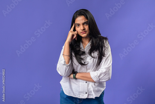 A young woman of Indian descent, wearing a white striped shirt and black smartwatch, poses thoughtfully with her finger on her temple, thought against a solid purple background