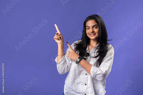 A young woman of Indian descent, wearing a white striped shirt and black smartwatch, poses confidently against a solid purple background, pointing to the side with her index finger.