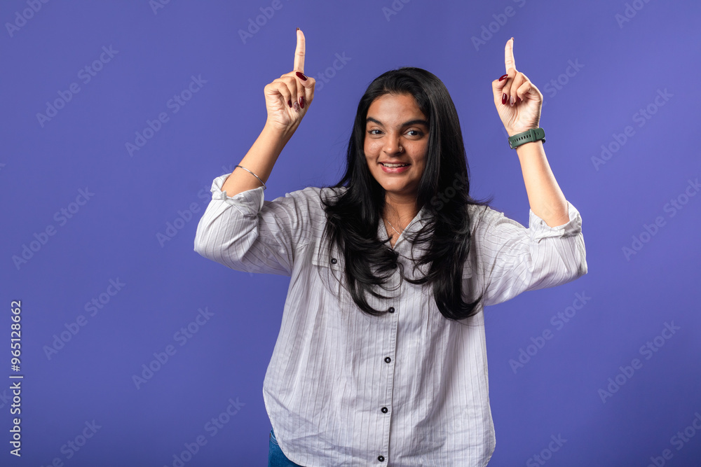 A young woman of Indian descent, wearing a white striped shirt and black smartwatch, poses confidently against a solid purple background, pointing to the side with her index finger. A young woman of Indian descent, wearing a white striped shirt and black smartwatch, poses confidently against a solid purple background, pointing to the side with her index finger.