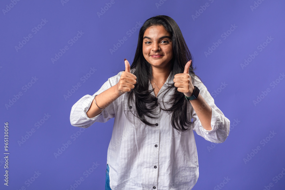 A young woman of Indian descent, wearing a white striped shirt and black smartwatch, stands against a solid purple background, smiling and giving thumbs-up gesture, radiating approval. A young woman of Indian descent, wearing a white striped shirt and black smartwatch, stands against a solid purple background, smiling and giving thumbs-up gesture, radiating approval.