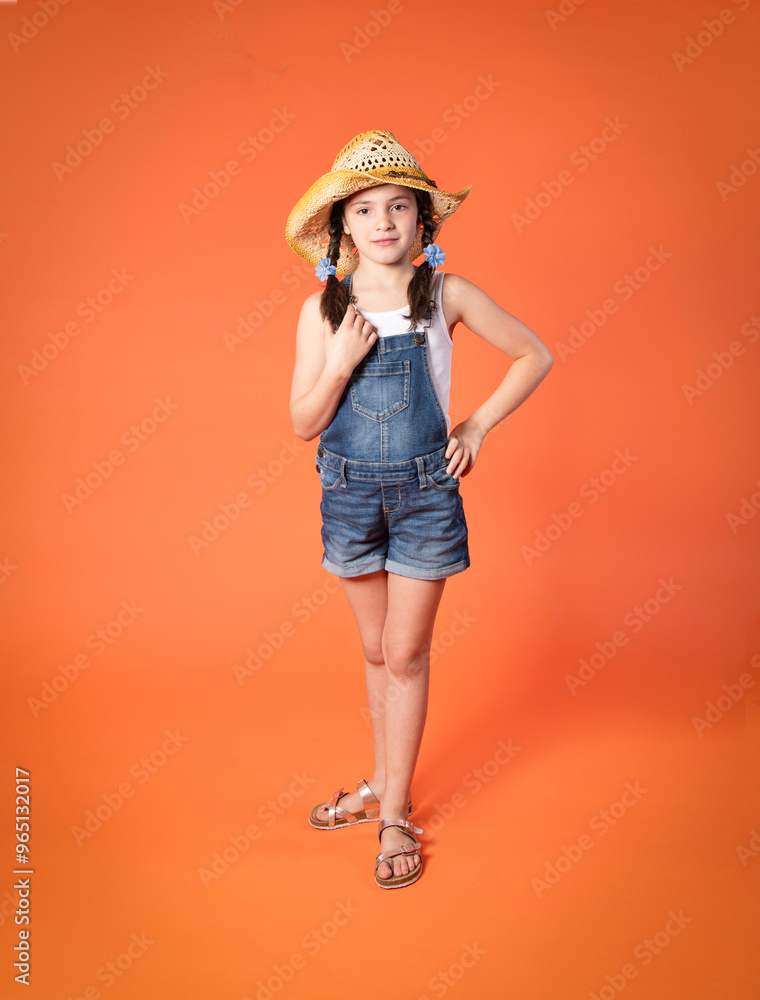 Adorable little girl in jean shorts and straw cowboy hat, standing isolated on orange background Adorable little girl in jean shorts and straw cowboy hat, standing isolated on orange background