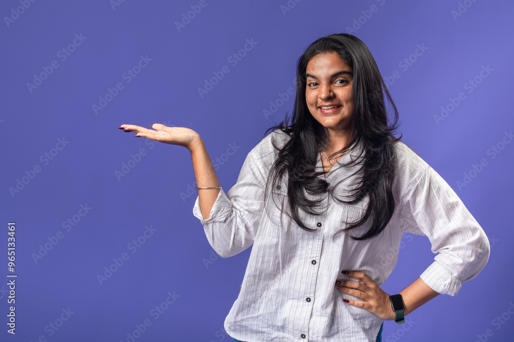 A young woman of Indian descent, wearing a white striped shirt and black smartwatch, stands against a solid purple background, smiling and gesturing with her palm up A young woman of Indian descent, wearing a white striped shirt and black smartwatch, stands against a solid purple background, smiling and gesturing with her palm up