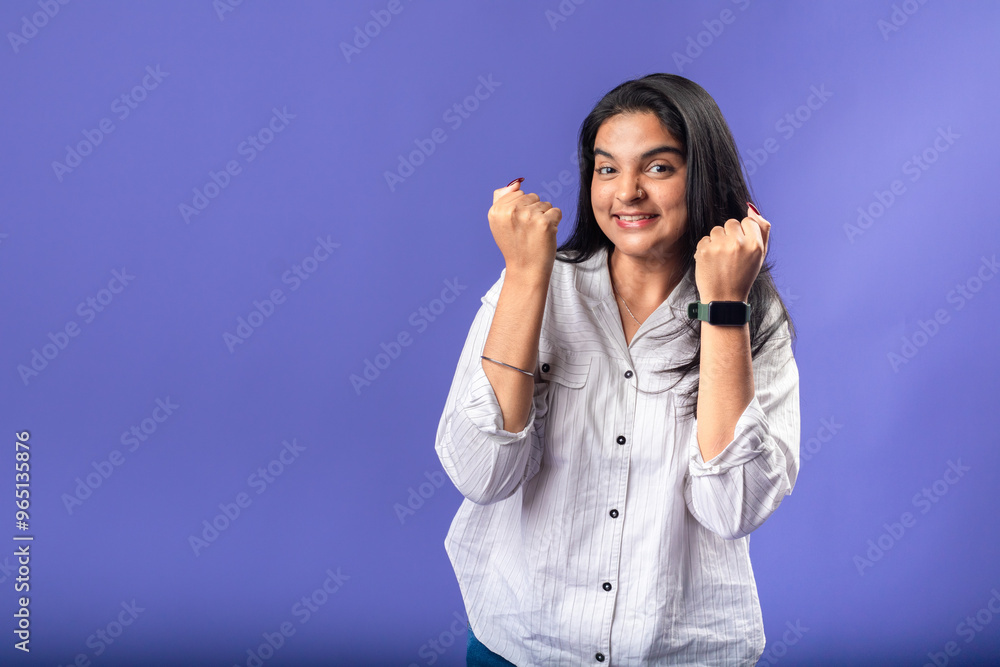 A young woman of Indian descent, wearing a white striped shirt and black smartwatch, stands against a solid purple background with her arms raised in a victory pose celebrating success A young woman of Indian descent, wearing a white striped shirt and black smartwatch, stands against a solid purple background with her arms raised in a victory pose celebrating success