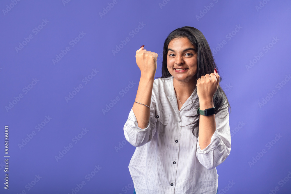 A young woman of Indian descent, wearing a white striped shirt and black smartwatch, stands against a solid purple background with her arms raised in a victory pose celebrating success A young woman of Indian descent, wearing a white striped shirt and black smartwatch, stands against a solid purple background with her arms raised in a victory pose celebrating success
