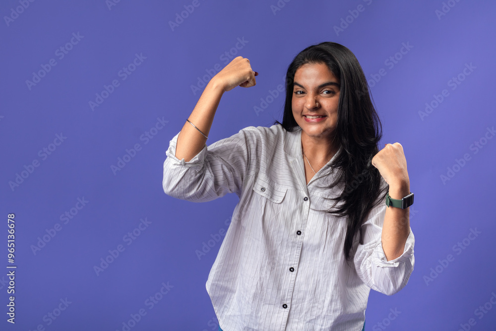 A young woman of Indian descent, wearing a white striped shirt and black smartwatch, stands against a solid purple background with her arms raised in a victory pose celebrating success A young woman of Indian descent, wearing a white striped shirt and black smartwatch, stands against a solid purple background with her arms raised in a victory pose celebrating success