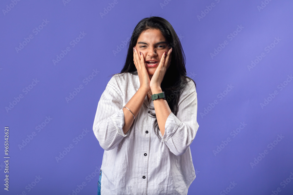 A young woman of Indian descent, wearing a white striped shirt and black smartwatch, stands against a solid purple background, with her hands on her cheeks, expressing surprise A young woman of Indian descent, wearing a white striped shirt and black smartwatch, stands against a solid purple background, with her hands on her cheeks, expressing surprise