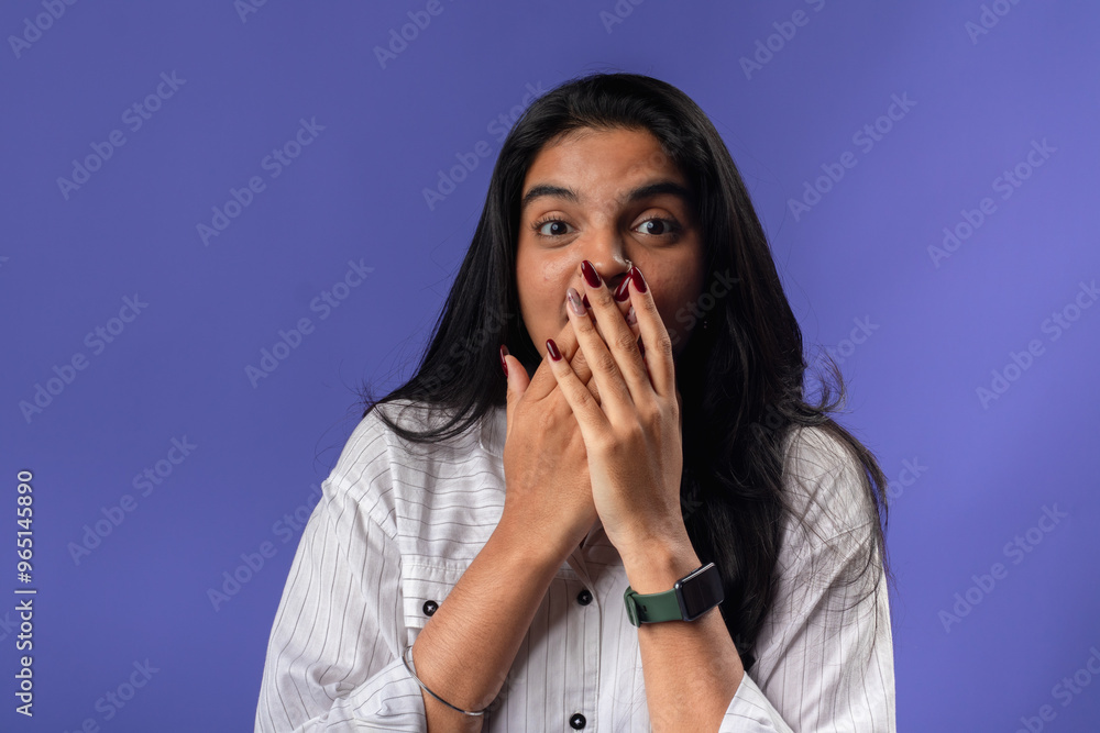 A young woman of Indian descent, wearing a white striped shirt and black smartwatch, stands against a solid purple background, covering her mouth with both hands, expressing surprise A young woman of Indian descent, wearing a white striped shirt and black smartwatch, stands against a solid purple background, covering her mouth with both hands, expressing surprise