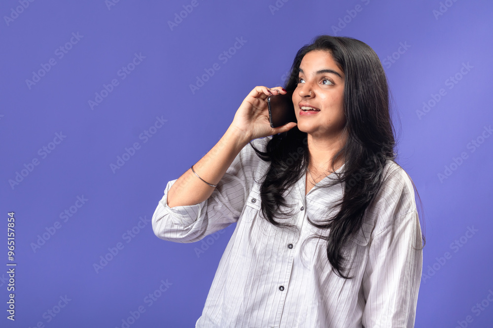 A young woman of Indian, wearing a white striped shirt and black smartwatch, against a solid purple background, holding a smartphone to her ear, as if having a phone conversation. A young woman of Indian, wearing a white striped shirt and black smartwatch, against a solid purple background, holding a smartphone to her ear, as if having a phone conversation.