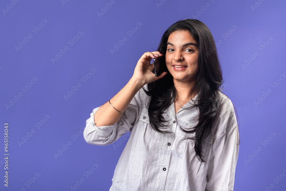A young woman of Indian, wearing a white striped shirt and black smartwatch, against a solid purple background, holding a smartphone to her ear, as if having a phone conversation. A young woman of Indian, wearing a white striped shirt and black smartwatch, against a solid purple background, holding a smartphone to her ear, as if having a phone conversation.