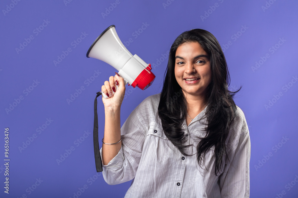 A young woman of Indian descent, wearing a white striped shirt and black smartwatch, stands against a solid purple background, smiling and holding a megaphone, suggesting communication. A young woman of Indian descent, wearing a white striped shirt and black smartwatch, stands against a solid purple background, smiling and holding a megaphone, suggesting communication.