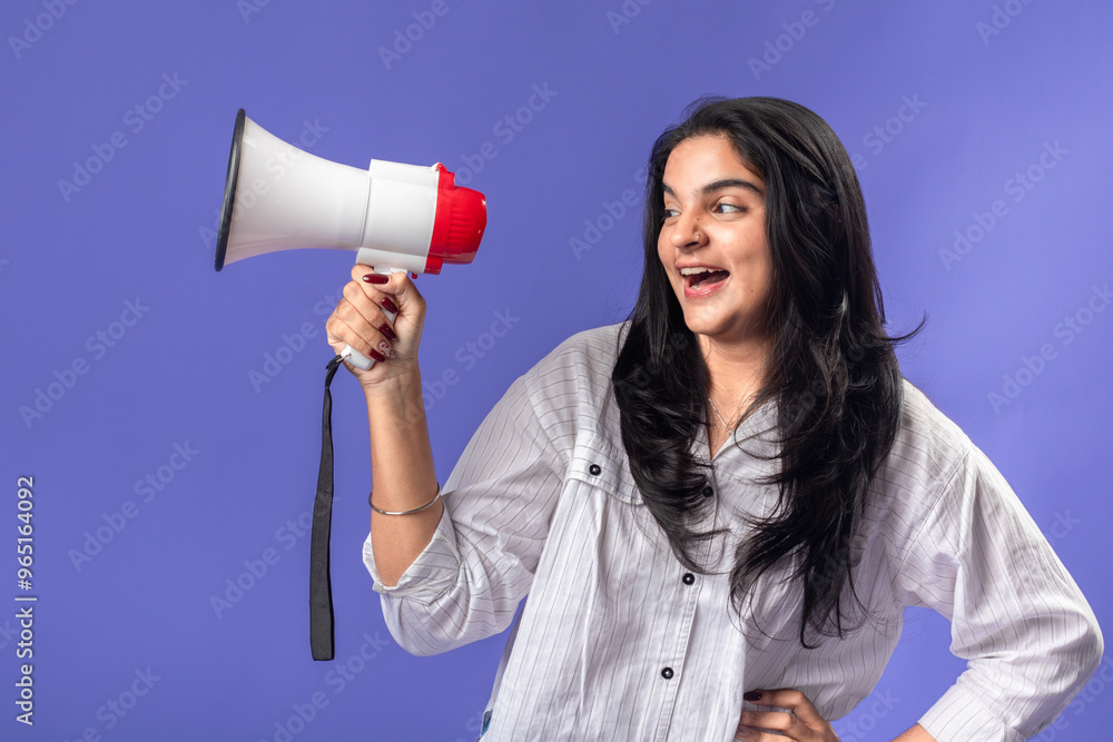 A young woman of Indian descent, wearing a white striped shirt and black smartwatch, stands against a solid purple background, smiling and holding a megaphone, suggesting communication. A young woman of Indian descent, wearing a white striped shirt and black smartwatch, stands against a solid purple background, smiling and holding a megaphone, suggesting communication.