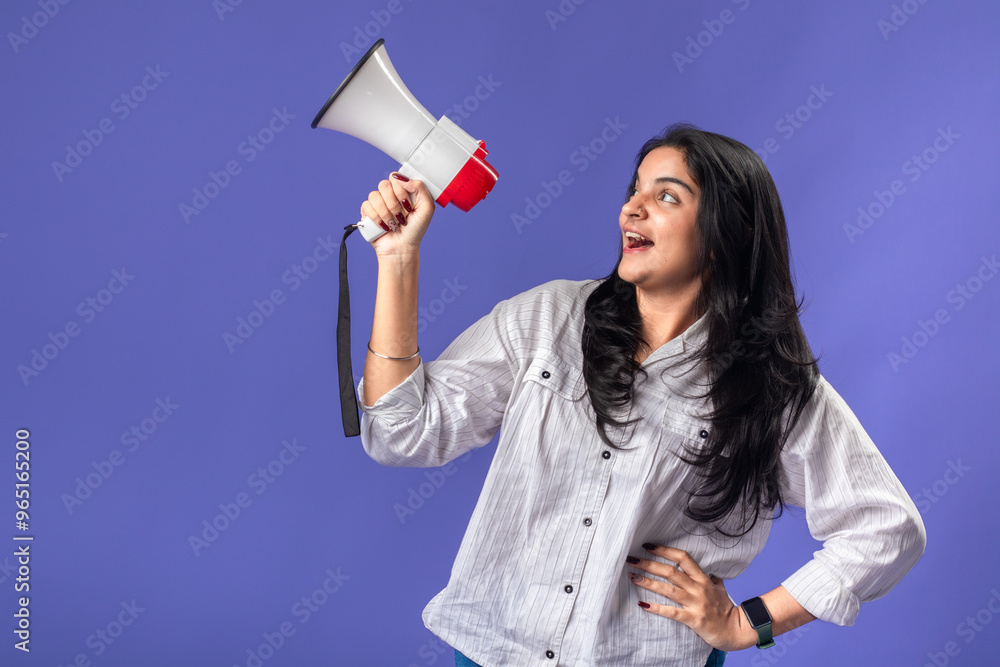 A young woman of Indian descent, wearing a white striped shirt and black smartwatch, stands against a solid purple background, smiling and holding a megaphone, suggesting communication. A young woman of Indian descent, wearing a white striped shirt and black smartwatch, stands against a solid purple background, smiling and holding a megaphone, suggesting communication.