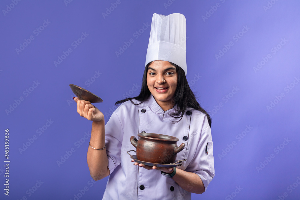 An Indian woman wearing a purple chef coat and white chef hat is smiling while holding a clay pot in one hand and its lid in the other. stands against a purple background An Indian woman wearing a purple chef coat and white chef hat is smiling while holding a clay pot in one hand and its lid in the other. stands against a purple background