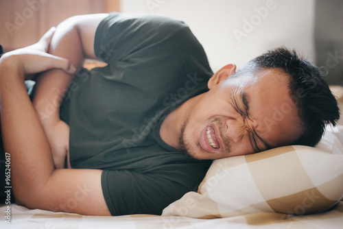 An Asian man lying in bed holding his stomach with a pained expression diarrhea, gerd concept