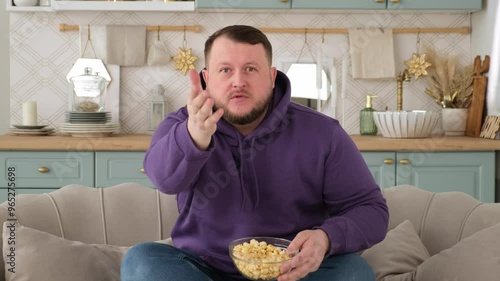 man of 30-35 years old in a purple hoodie sits on the sofa in front of the TV, is indignant and swears