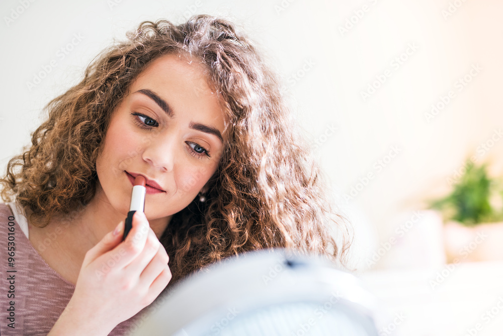 Young woman sitting on bed, applying red lipstick to her lips, looking into a round makeup mirror. Young woman sitting on bed, applying red lipstick to her lips, looking into a round makeup mirror.