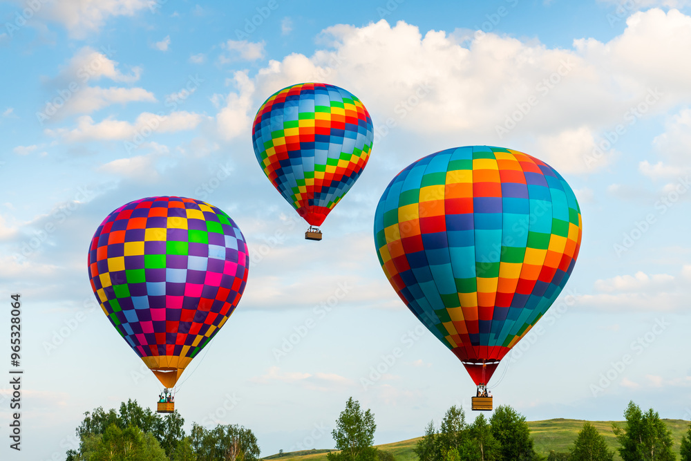 Colorful hot air balloons flying over the green hills and trees at sunset. Rainbow colored balloons in the blue sky.