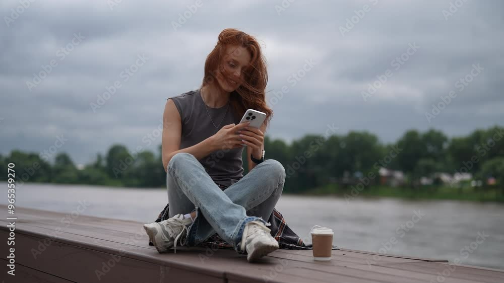 Happy young woman enjoying peaceful moment sitting on wooden pier by river, smiling using smartphone to savor break in urban nature on cloudy summer day. Serene female enjoying free time outdoors. Happy young woman enjoying peaceful moment sitting on wooden pier by river, smiling using smartphone to savor break in urban nature on cloudy summer day. Serene female enjoying free time outdoors.