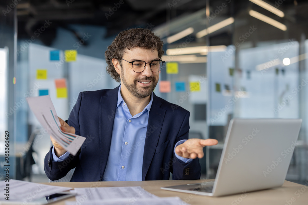 Businessman in office holding reports while engaging in discussion using laptop. Scene captures corporate environment with focus on analysis and communication Businessman in office holding reports while engaging in discussion using laptop. Scene captures corporate environment with focus on analysis and communication